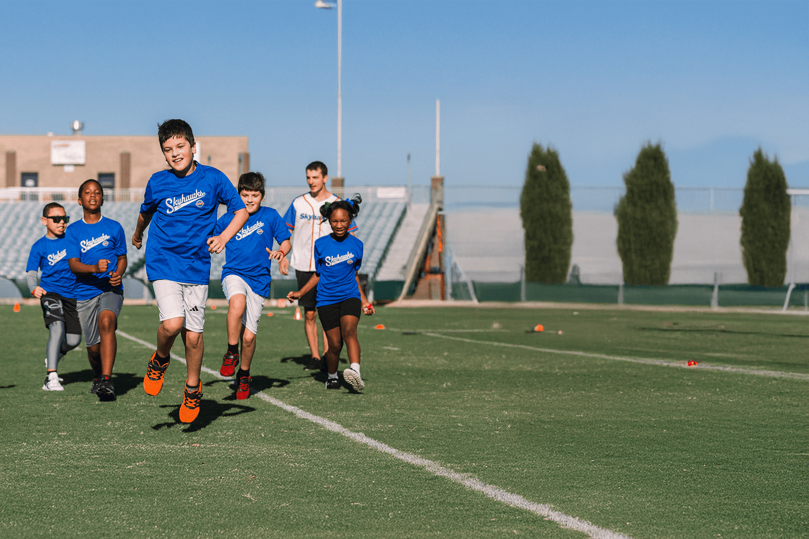 A group of children in blue shirts dash across a grassy field under the Colorado Springs sun, with a building and trees providing a picturesque backdrop. - Skyhawks-Supertots Colorado