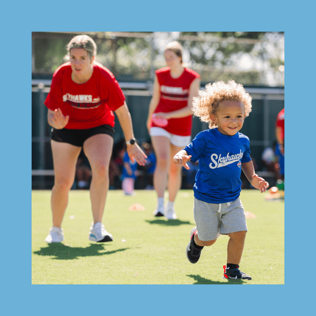 A child in a blue shirt joyfully sprints across the grass, while adults in red shirts, possibly Skyhawks coaches or SuperTots Sports instructors, watch attentively in the background. This lively scene could easily be taking place on one of Colorado's scenic fields. - Skyhawks-Supertots Colorado