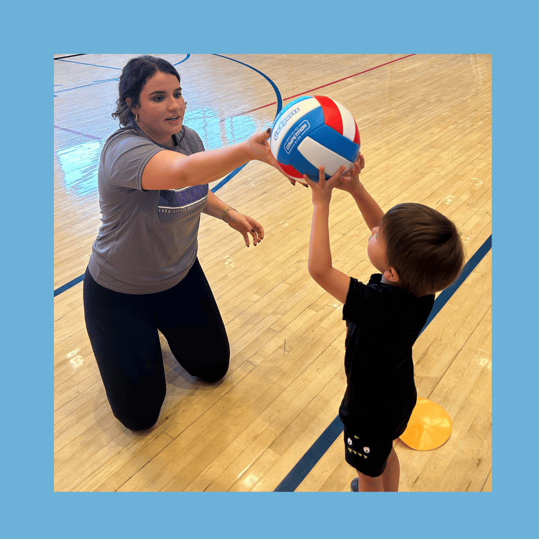 Adult kneels, handing colorful volleyball to child in gym. Child stands, reaching out. Blue framed image captures the joy of learning with SuperTots Sports in Colorado. - Skyhawks-Supertots Colorado