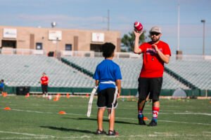 A Skyhawks coach in a red shirt and hat holds a football and talks to a young player on a field with empty bleachers in the background. - Skyhawks-Supertots Colorado