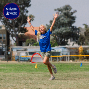 A young girl in a blue SuperTots Sports shirt raises her arms and smiles while running on a grassy sports field, with cones and trees in the background—ready to get Back in Action this season!. - Skyhawks-Supertots Colorado