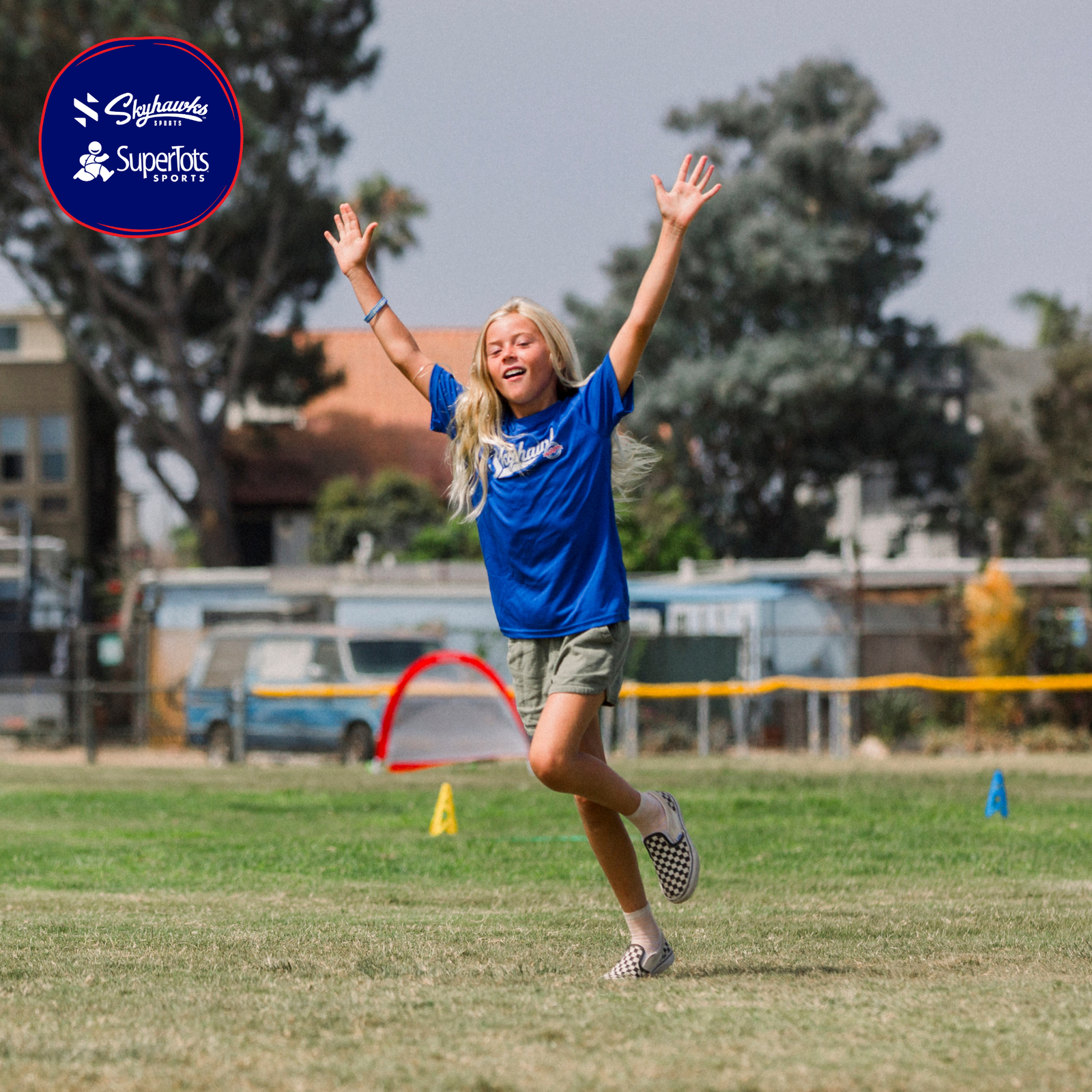 A young girl in a blue SuperTots Sports shirt raises her arms and smiles while running on a grassy sports field, with cones and trees in the background—ready to get Back in Action this season!. - Skyhawks-Supertots Colorado
