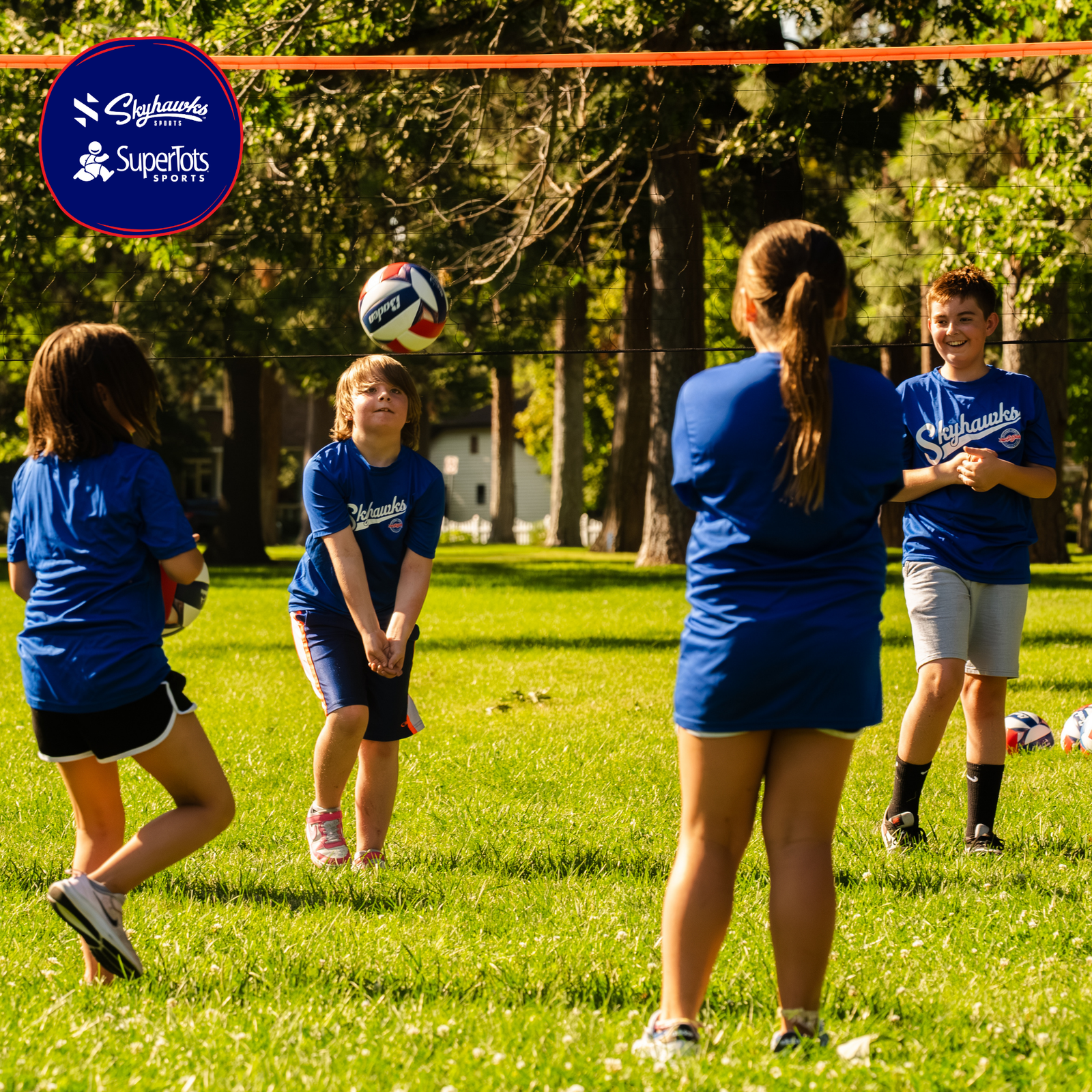 Four kids in blue shirts play volleyball outside on grass under trees, with a net above them—building character and teamwork on the field. A logo in the top left corner reads "Skyhawks SuperTots Sports. - Skyhawks-Supertots Colorado