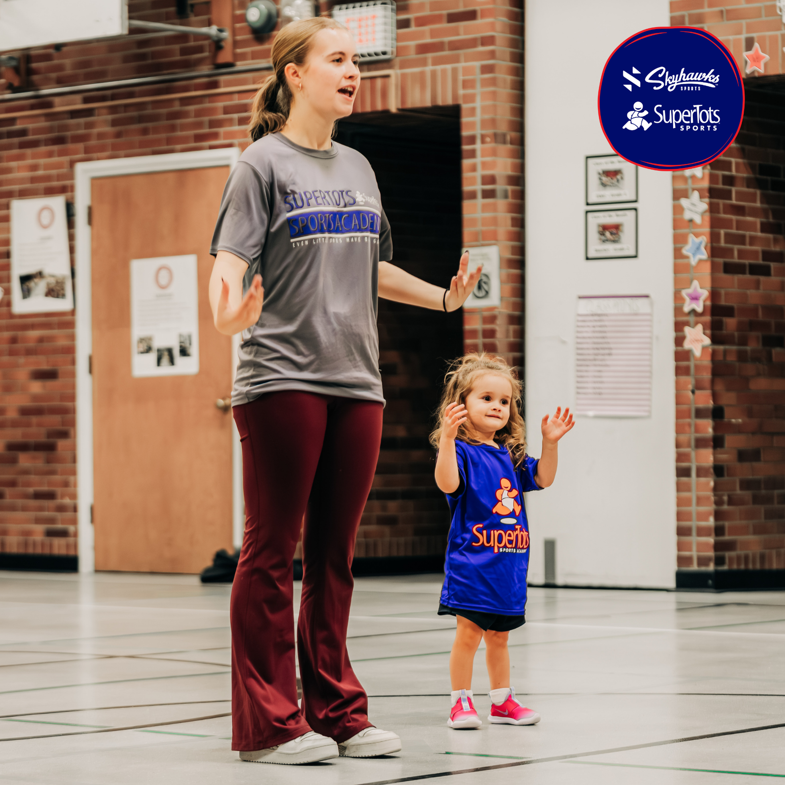 An adult and a young child stand in a gym. The adult wears a gray shirt and maroon pants, while the child, dressed in a blue SuperTots shirt and pink shoes, represents Building Character on the Field. The SuperTots Sports logo is visible. - Skyhawks-Supertots Colorado