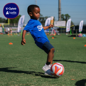 A young child in a blue shirt kicks a soccer ball on a grassy field during a Back to School sports activity. Sports equipment and banners are visible in the background, capturing the excitement of being back in action. - Skyhawks-Supertots Colorado