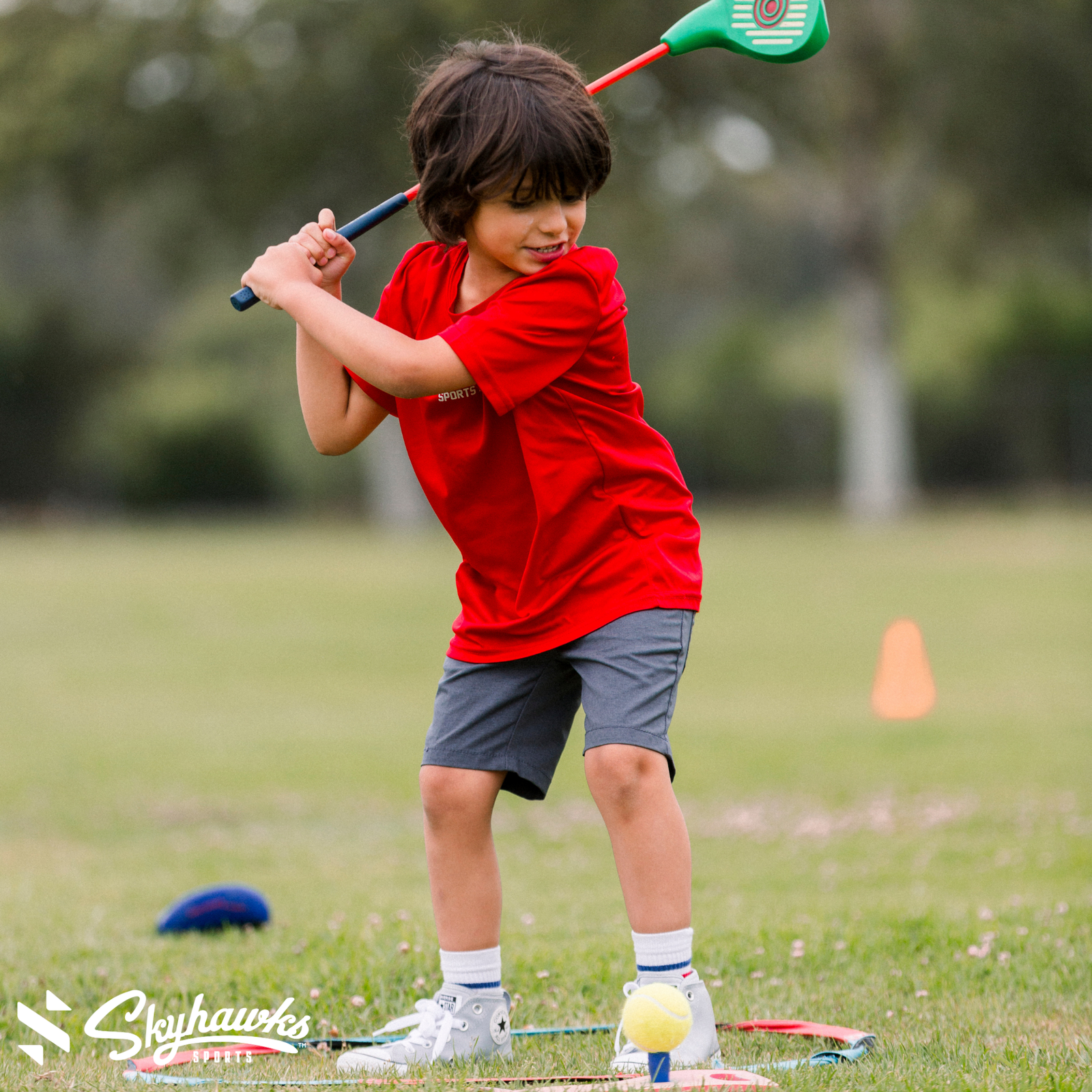 A young boy in a red shirt and gray shorts gets Back in Action as he prepares to hit a tennis ball with a plastic golf club on a grassy field. - Skyhawks-Supertots Colorado