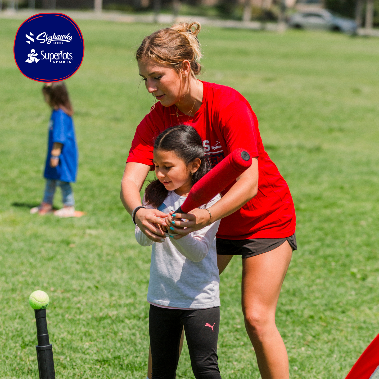 An adult helps a child hold a bat to hit a ball on a tee during a Back in Action sports activity on a grassy field. Two logos are visible in the top left corner. - Skyhawks-Supertots Colorado