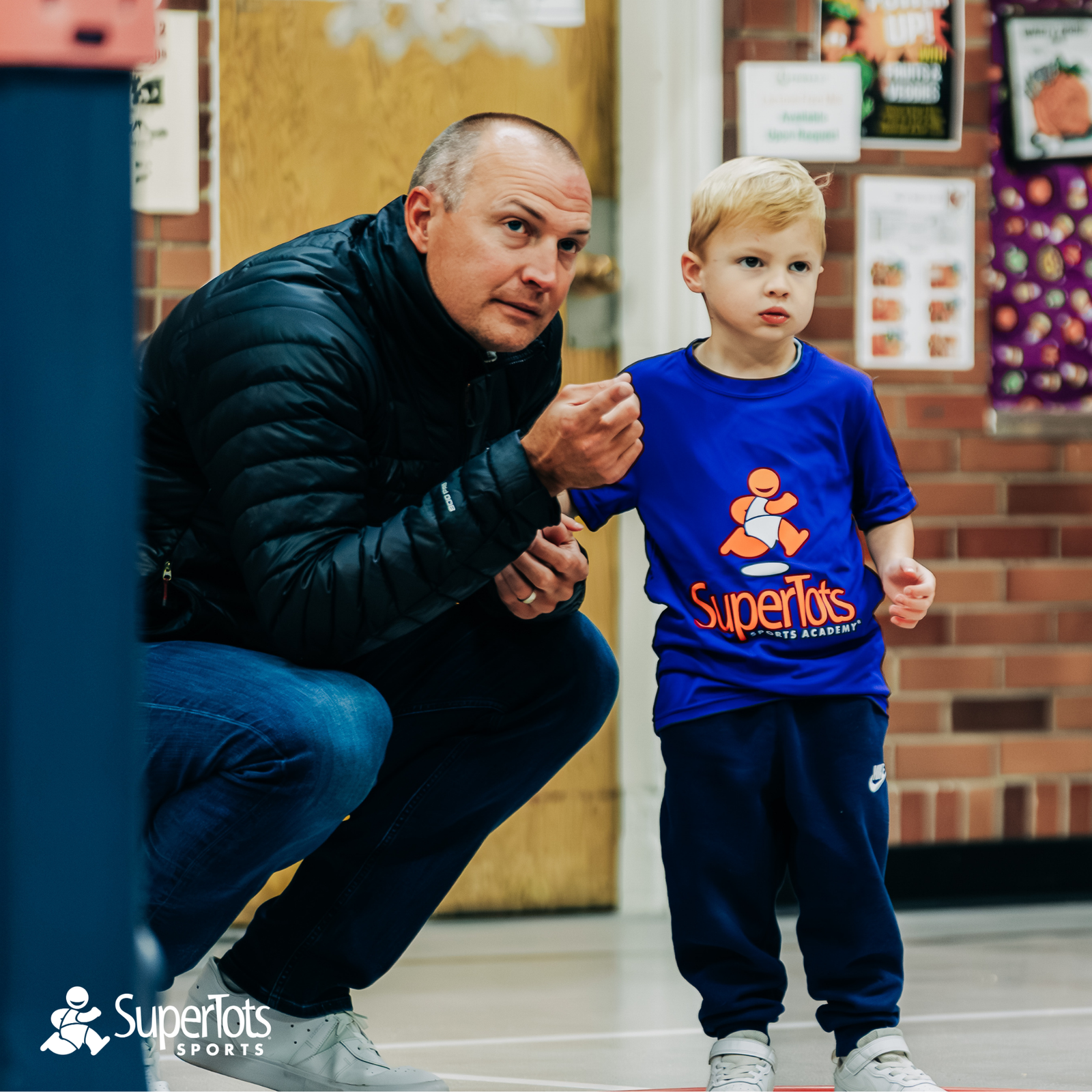 An adult crouches next to a young child in a SuperTots Sports shirt inside a gym, both focused on something out of frame—capturing a special moment in youth sports. The SuperTots Sports logo is visible at the bottom left. - Skyhawks-Supertots Colorado