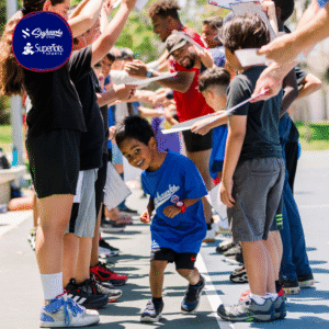 Young boy in a blue sports shirt runs through a tunnel of cheering people holding up hands and papers on an outdoor court. - Skyhawks-Supertots Colorado