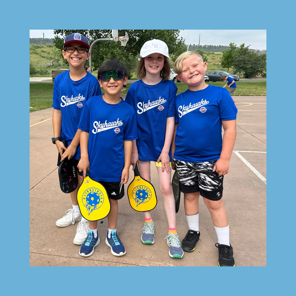 Four children in matching blue Skyhawks shirts stand outdoors on a basketball court in Colorado, holding yellow pickleball paddles and smiling at the camera. - Skyhawks-Supertots Colorado