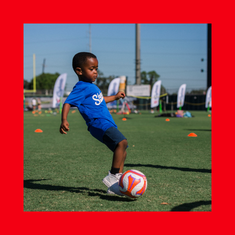 A young child in a blue shirt and shorts kicks a soccer ball on a grassy Colorado field marked with orange cones, enjoying SuperTots Sports activities. - Skyhawks-Supertots Colorado