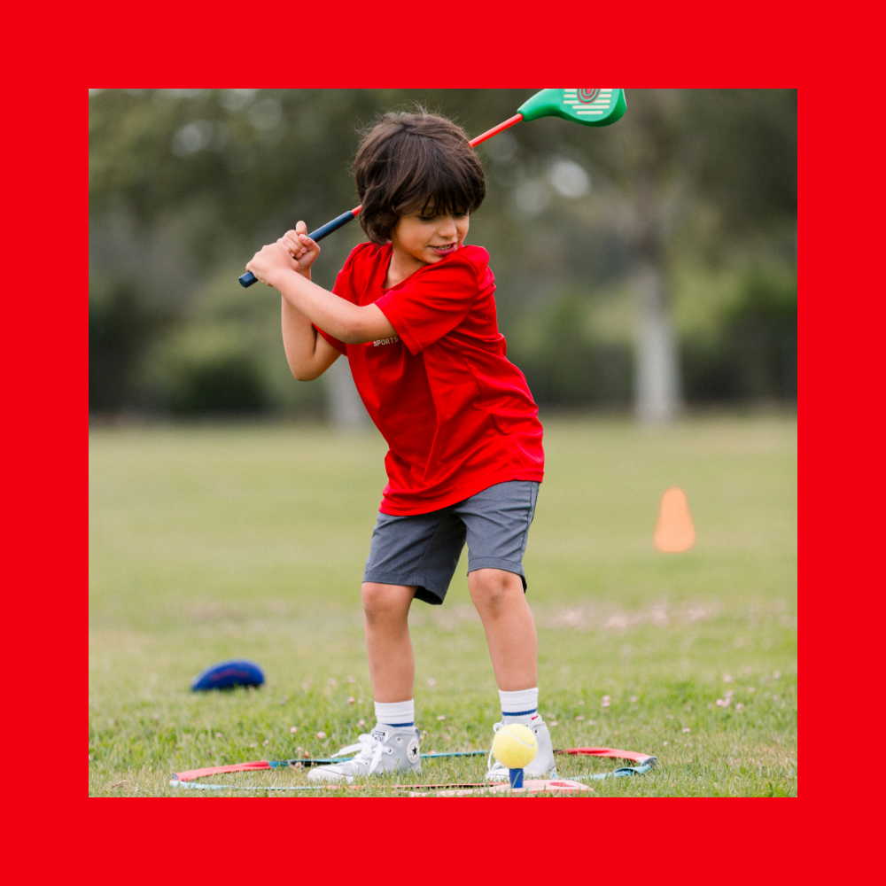 A young boy wearing a red shirt and gray shorts prepares to hit a plastic ball with a toy golf club on a grassy field at a SuperTots Sports event, part of Skyhawks Colorado, with cones visible in the background. - Skyhawks-Supertots Colorado