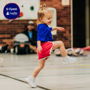A young child in athletic clothes is smiling and running indoors, lifting one leg off the ground, with sports equipment and adults in the background—a fun way to keep kids active during winter activities. - Skyhawks-Supertots Colorado