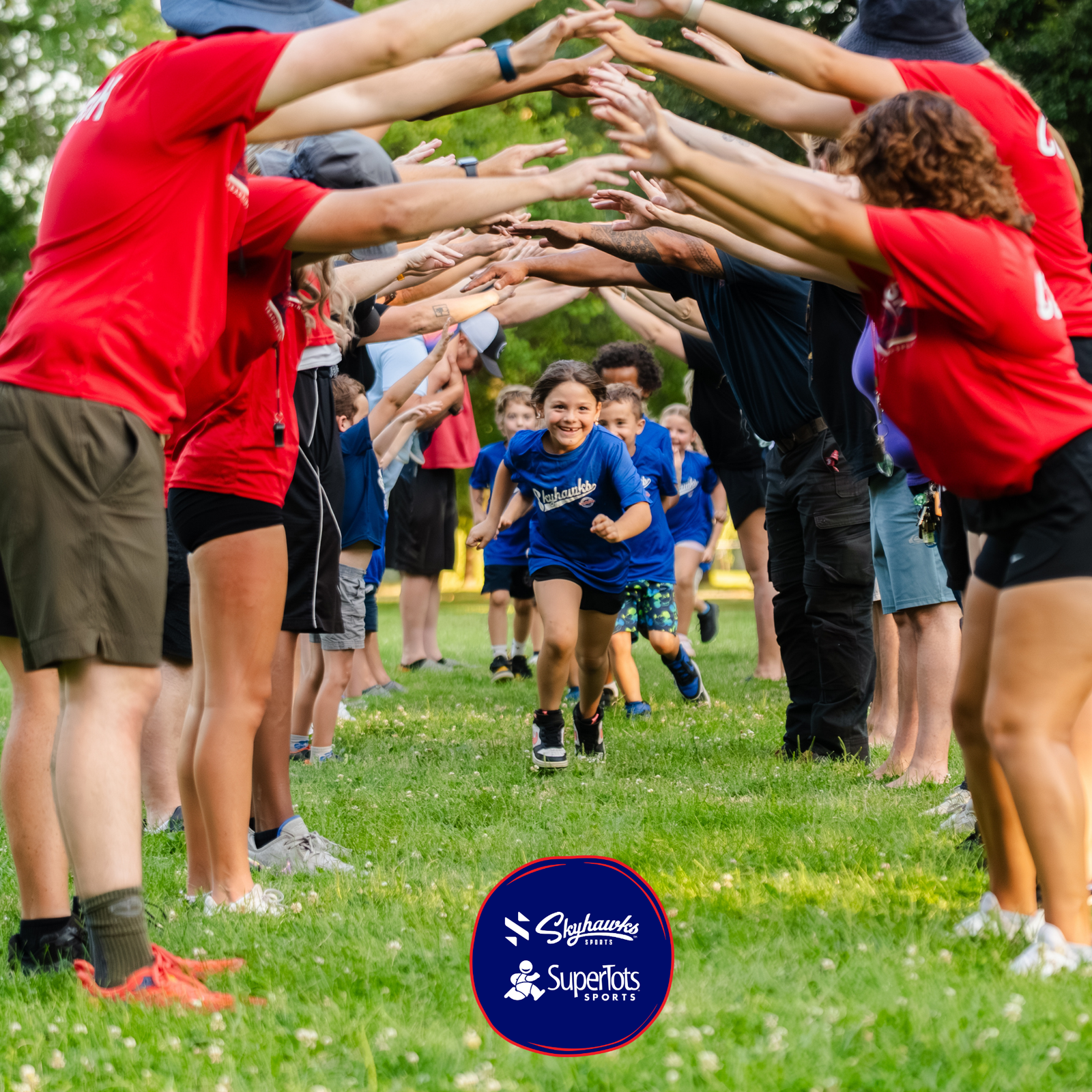 A child runs through a tunnel formed by adults and children holding up their arms at an outdoor event, showcasing camp for kids and the summer camp benefits offered by SuperTots and Skyhawks. - Skyhawks-Supertots Colorado