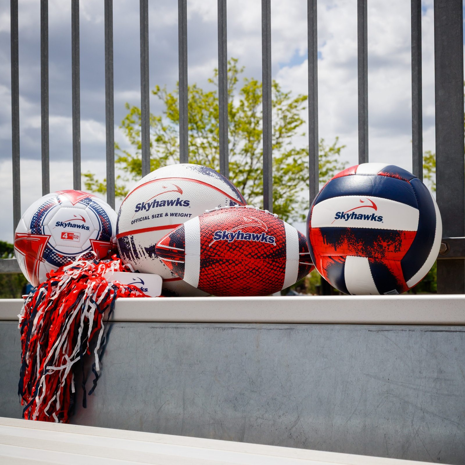 Four Skyhawks-branded sports balls and a red, white, and blue pom-pom rest on metal bleachers, with a fence and trees in the background—perfect for fans looking to cheer on their team or learn more about our discount process. - Skyhawks-Supertots Colorado