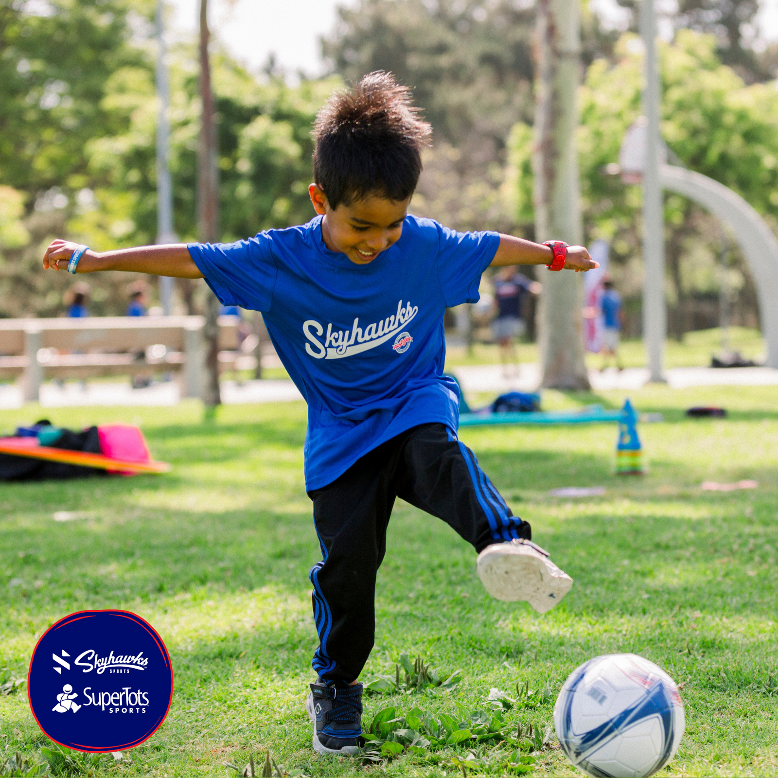 Young child in a blue "Skyhawks" t-shirt kicks a soccer ball on a grassy field during the day; SuperTots Sports logo is visible in the corner. - Skyhawks-Supertots Colorado