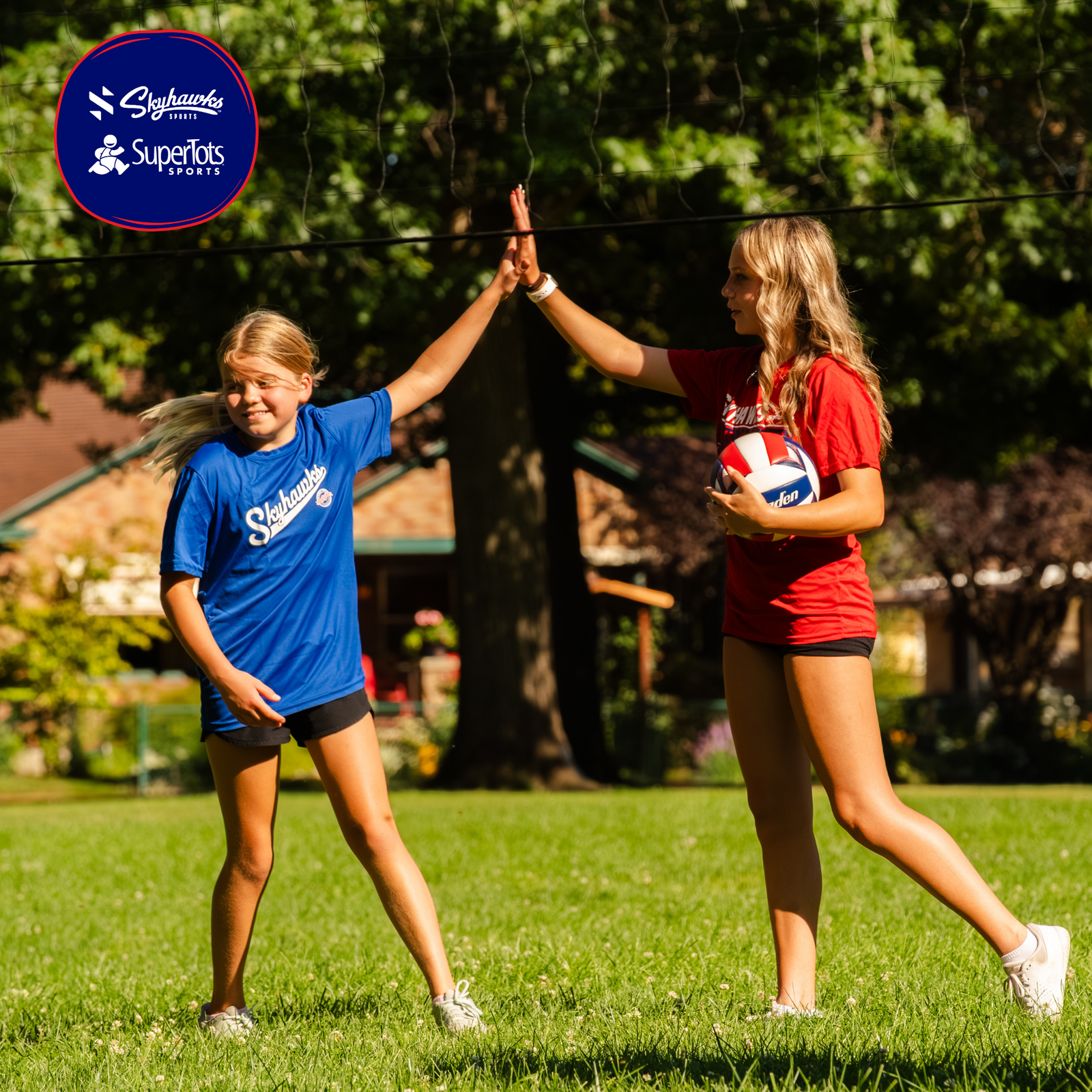 Two girls on a grassy field high-five each other; one holds a volleyball. Both wear Skyhawks and SuperTots Sports shirts, showing their passion for youth sports. Trees and a house are in the background. - Skyhawks-Supertots Colorado