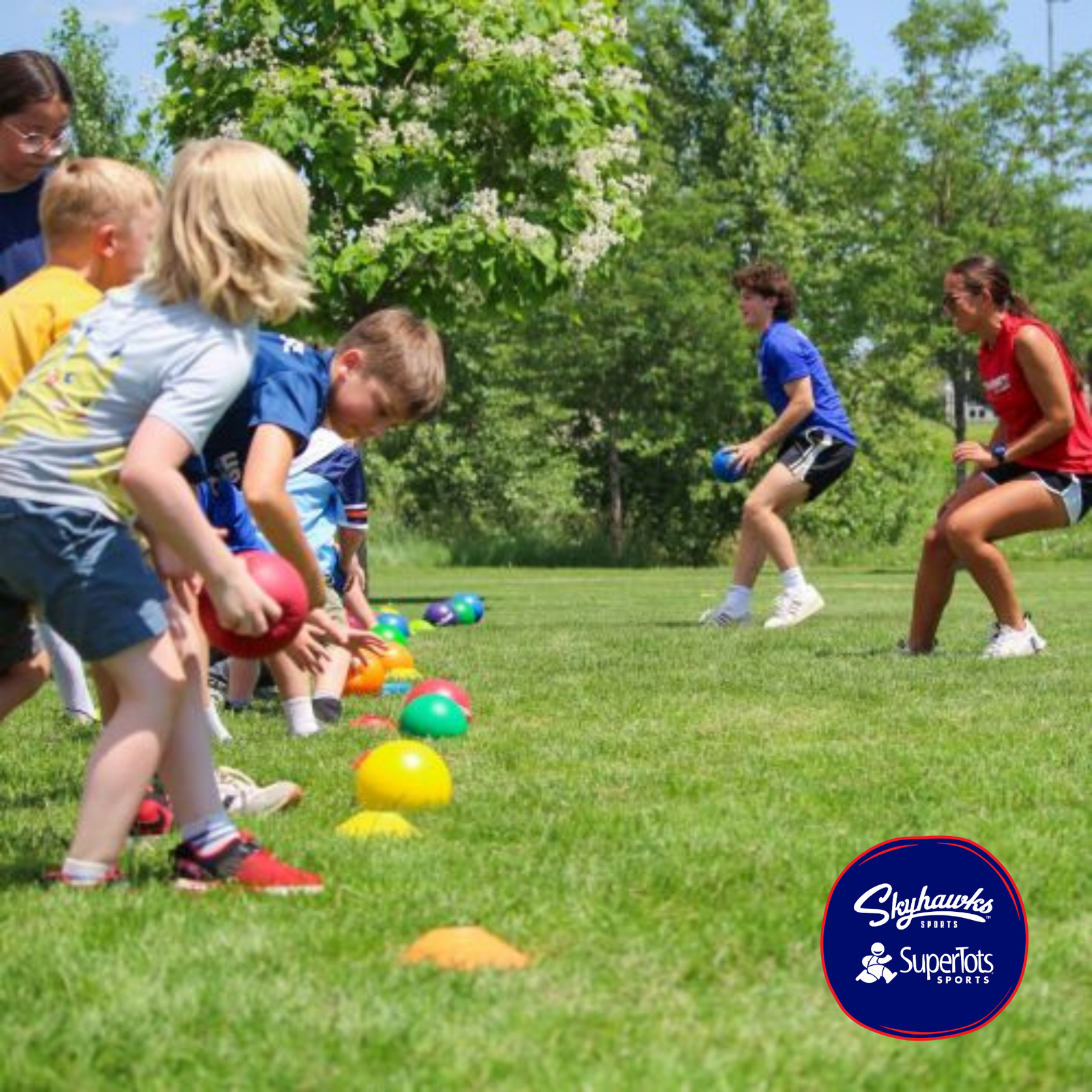 Children line up on grass holding balls, preparing for a sports activity outdoors, while two instructors watch. Engaging in sports helps kids build stronger brains and bodies. Trees and colorful cones are visible in the background. Skyhawks Sports logo is in the corner. - Skyhawks-Supertots Colorado