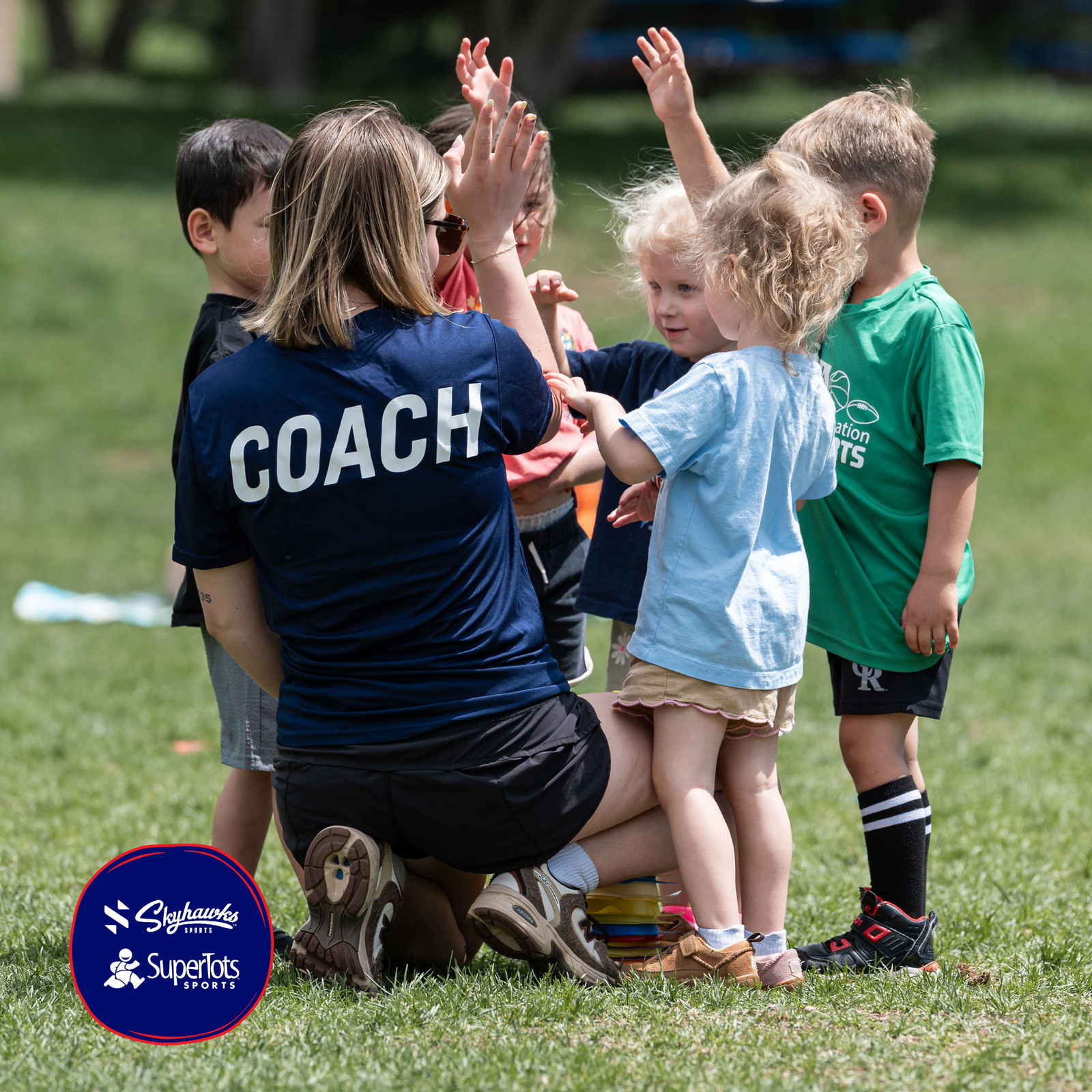 A coach kneels on grass, giving high-fives to a group of kids in sports attire during an outdoor activity that supports brain development. The SuperTots Sports logo is visible in the corner. - Skyhawks-Supertots Colorado