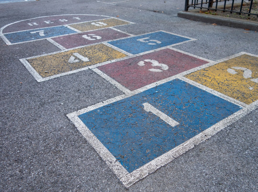 A hopscotch court is painted on asphalt, featuring numbered squares in blue, yellow, and red, with a semicircular “rest” area at the top—perfect for encouraging movement and helping children build confidence as they play. - Skyhawks-Supertots Colorado