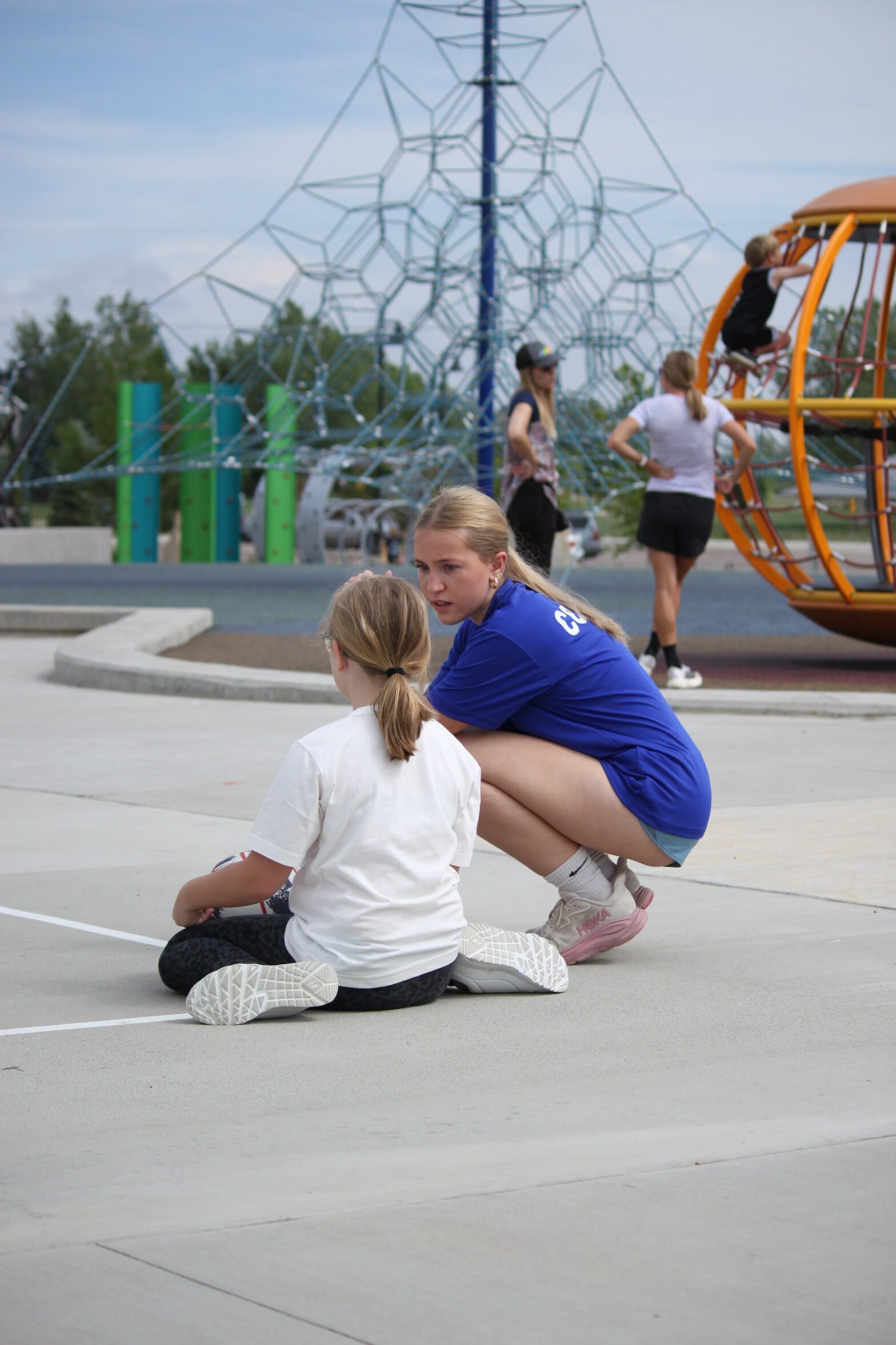 An older girl in a blue shirt crouches to mentor a younger girl sitting on the ground at a playground, guiding her through a learning process. - Skyhawks-Supertots Colorado
